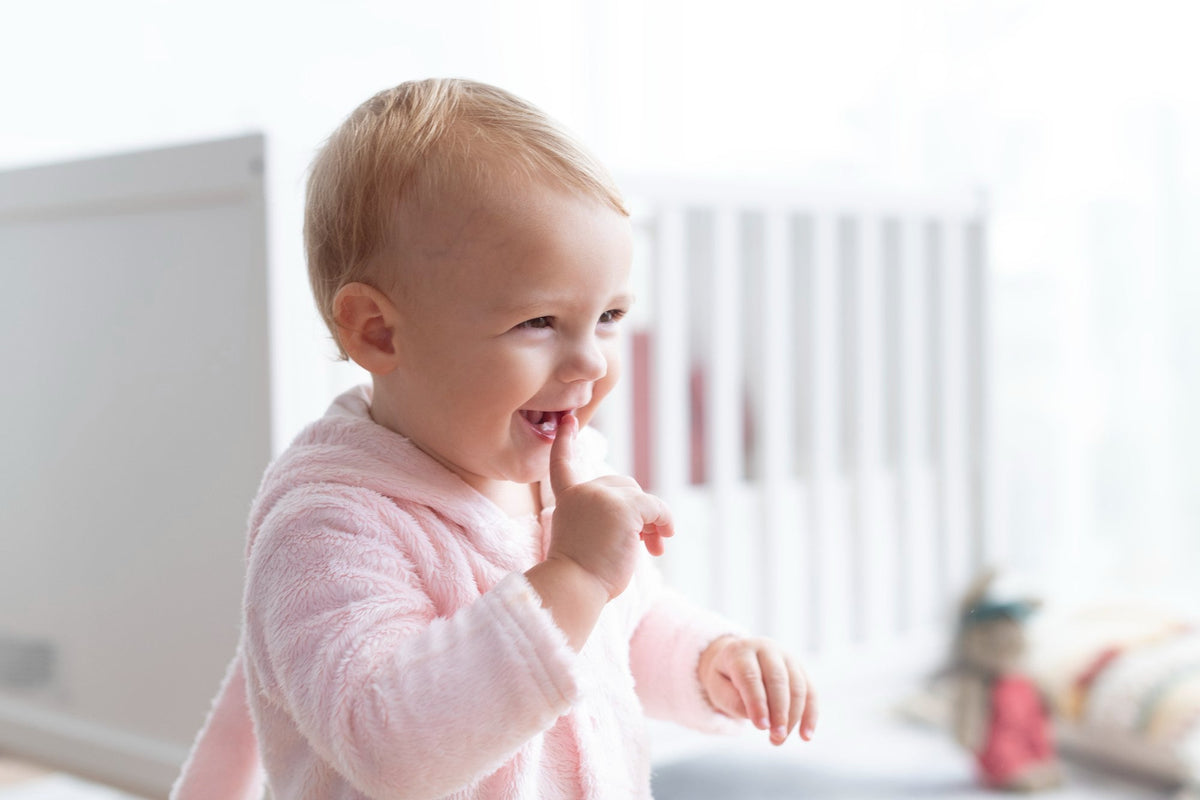 Smiling baby in a pink fleece dressing gown chewing on a finger, with a white cot behind