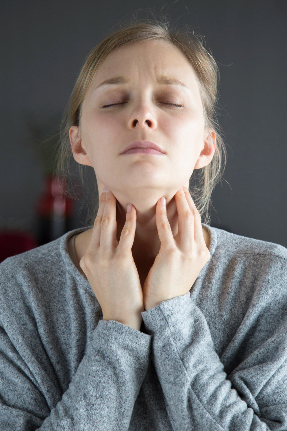 Young woman in a grey jumper with eyes closed, pressing her fingertips to either side of her sore throat