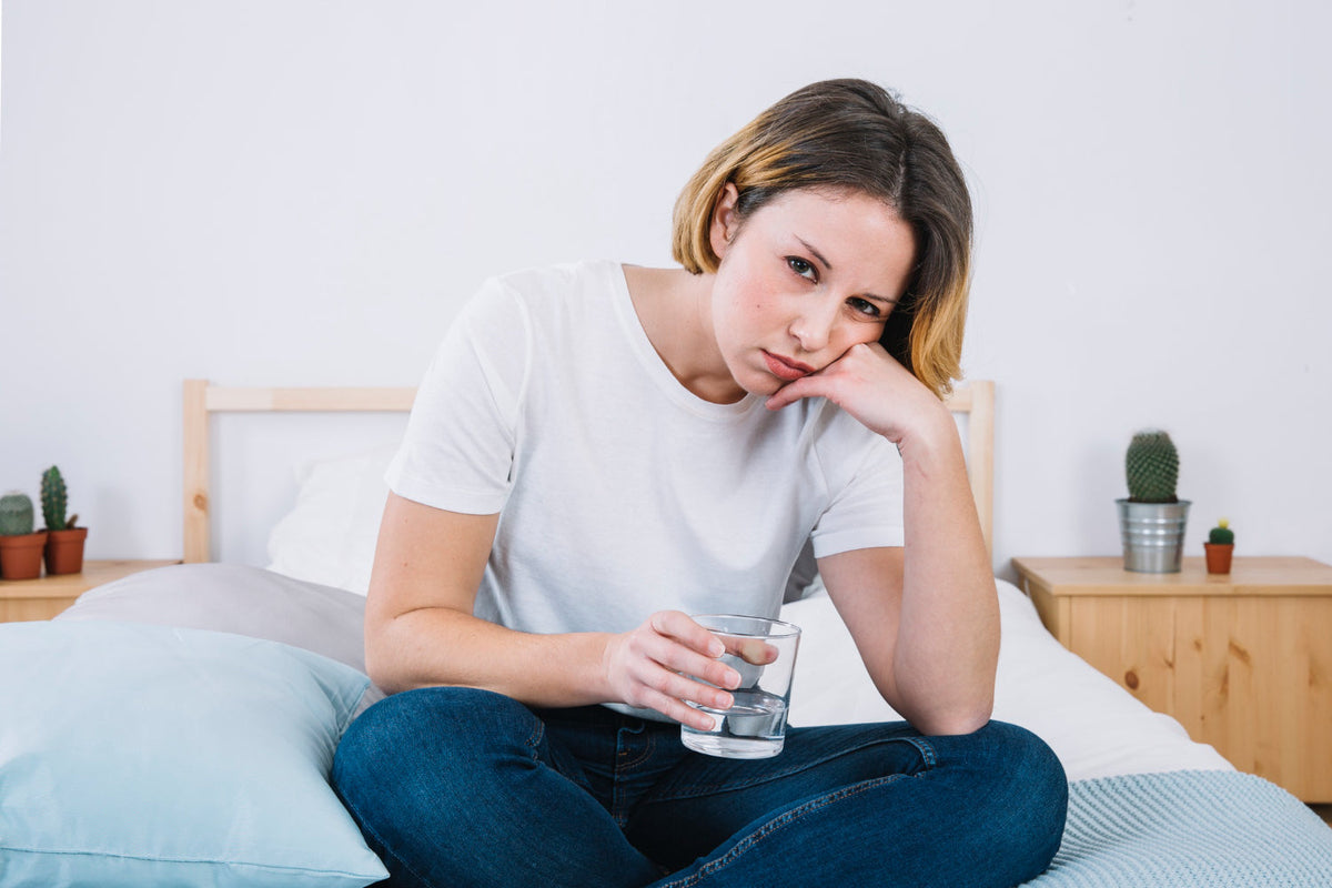 Tired-looking woman sitting on a bed, resting her head on her hand and holding a glass of water