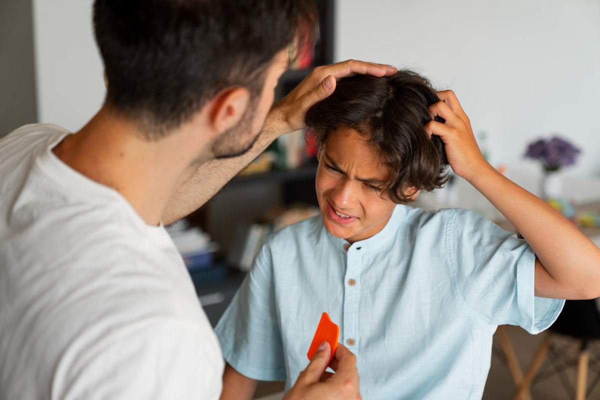 Man parting a grimacing boy's hair to check his scalp while holding a red fine-toothed comb