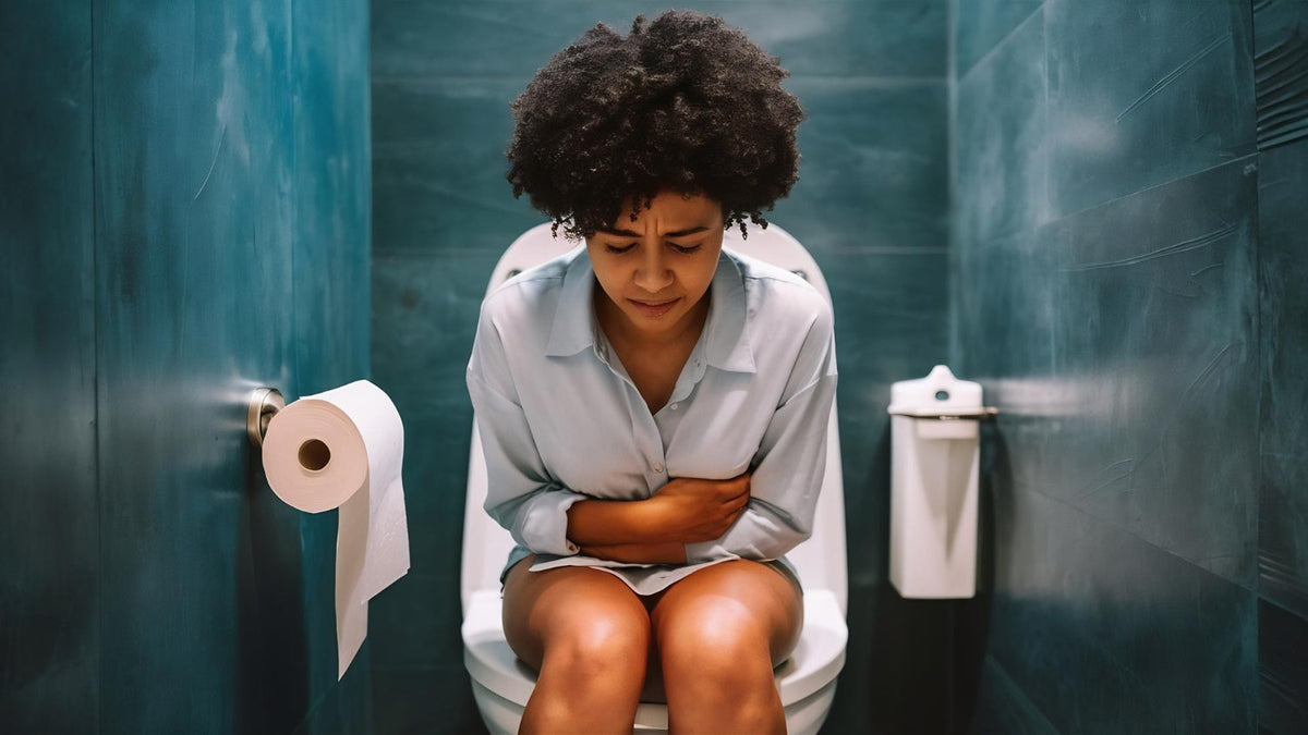 Woman sitting on a toilet in a teal-tiled bathroom, hunched forward and clutching her stomach