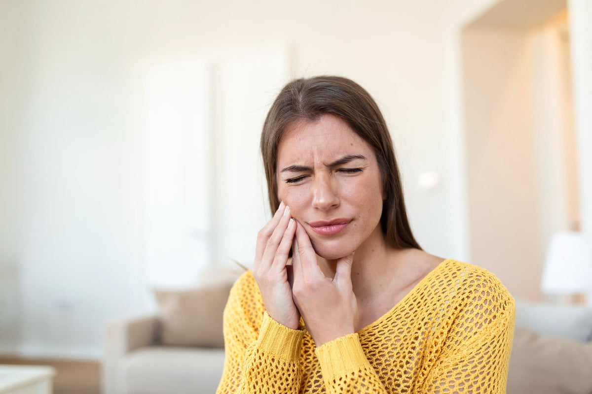 Woman in a yellow knitted jumper wincing and pressing her hand against her jaw in a living room