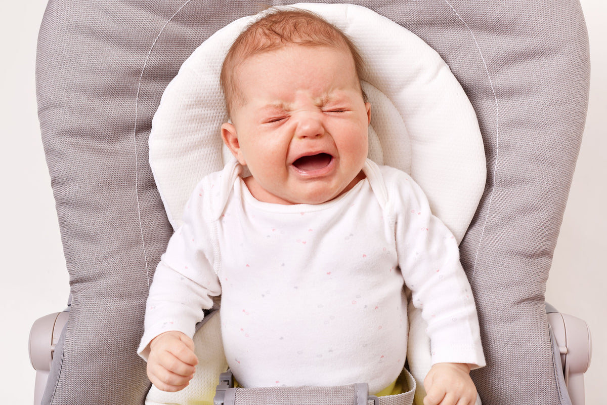 Baby in a white sleepsuit crying with eyes screwed shut while strapped into a grey bouncer chair