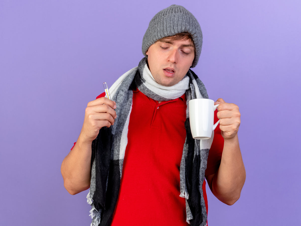 Unwell man in a winter hat and scarf holding a thermometer and a mug against a purple background